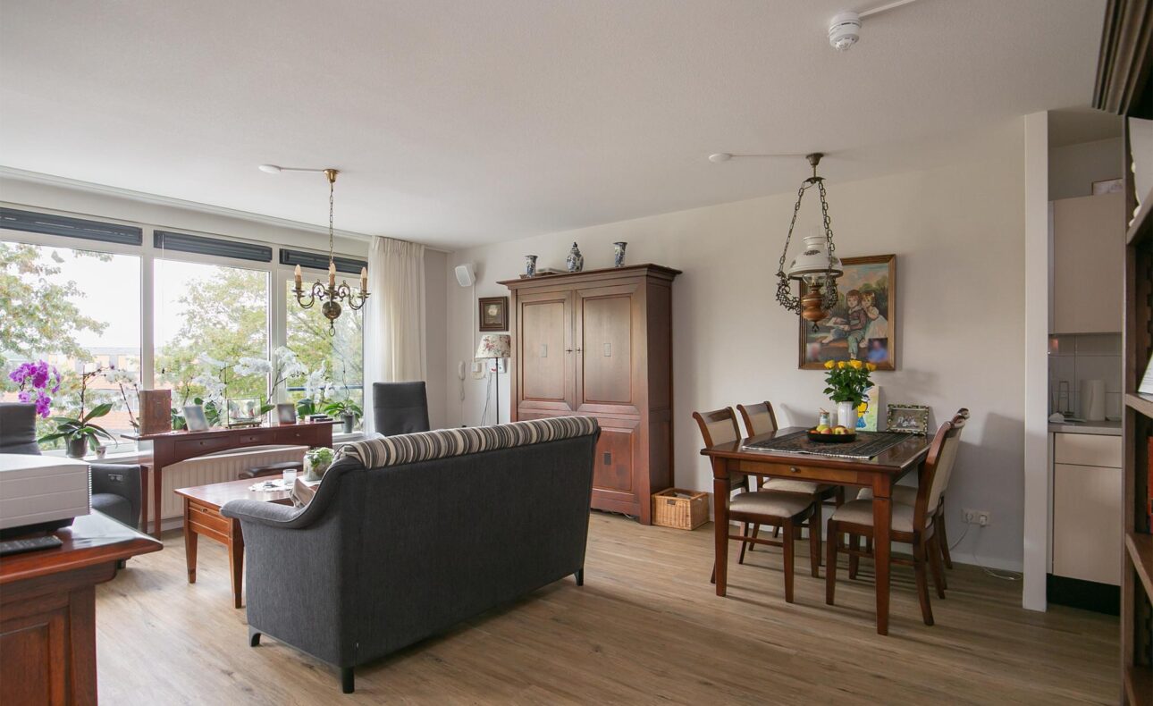 Woonkamer met houten vloer, grijze bank, eettafel met bloemen, grote kast en ramen met uitzicht op groene bomen.