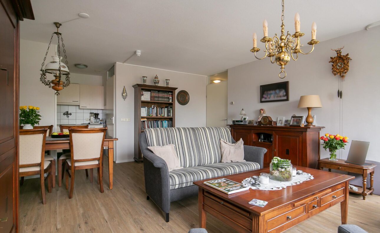 Woonkamer met klassieke inrichting, houten meubels, een bank, kroonluchter, en een eettafel. Planten en decoraties aanwezig.