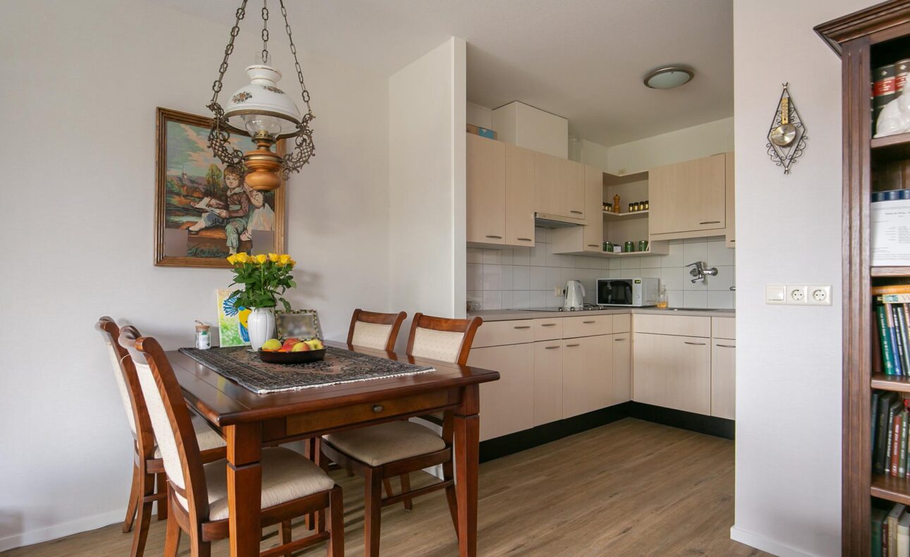 Eetkamer met houten tafel en vier stoelen, naast een keuken met lichte kasten. Gele bloemen in een vaas op tafel.