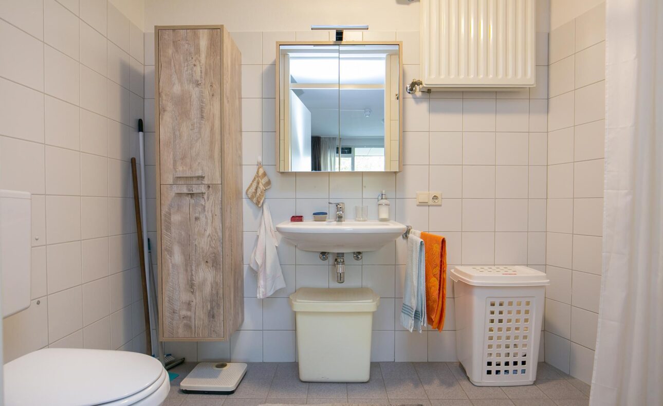 Badkamer met witte tegels, wastafel met spiegel, houten kast, wasmand en toilet. Handdoeken hangen aan de wastafel.