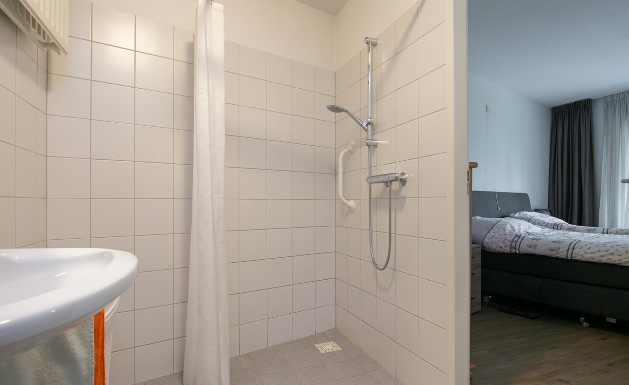Afbeelding van een moderne badkamer met een inloopdouche, witte tegels en een douchegordijn. Op de achtergrond is een slaapkamer zichtbaar.