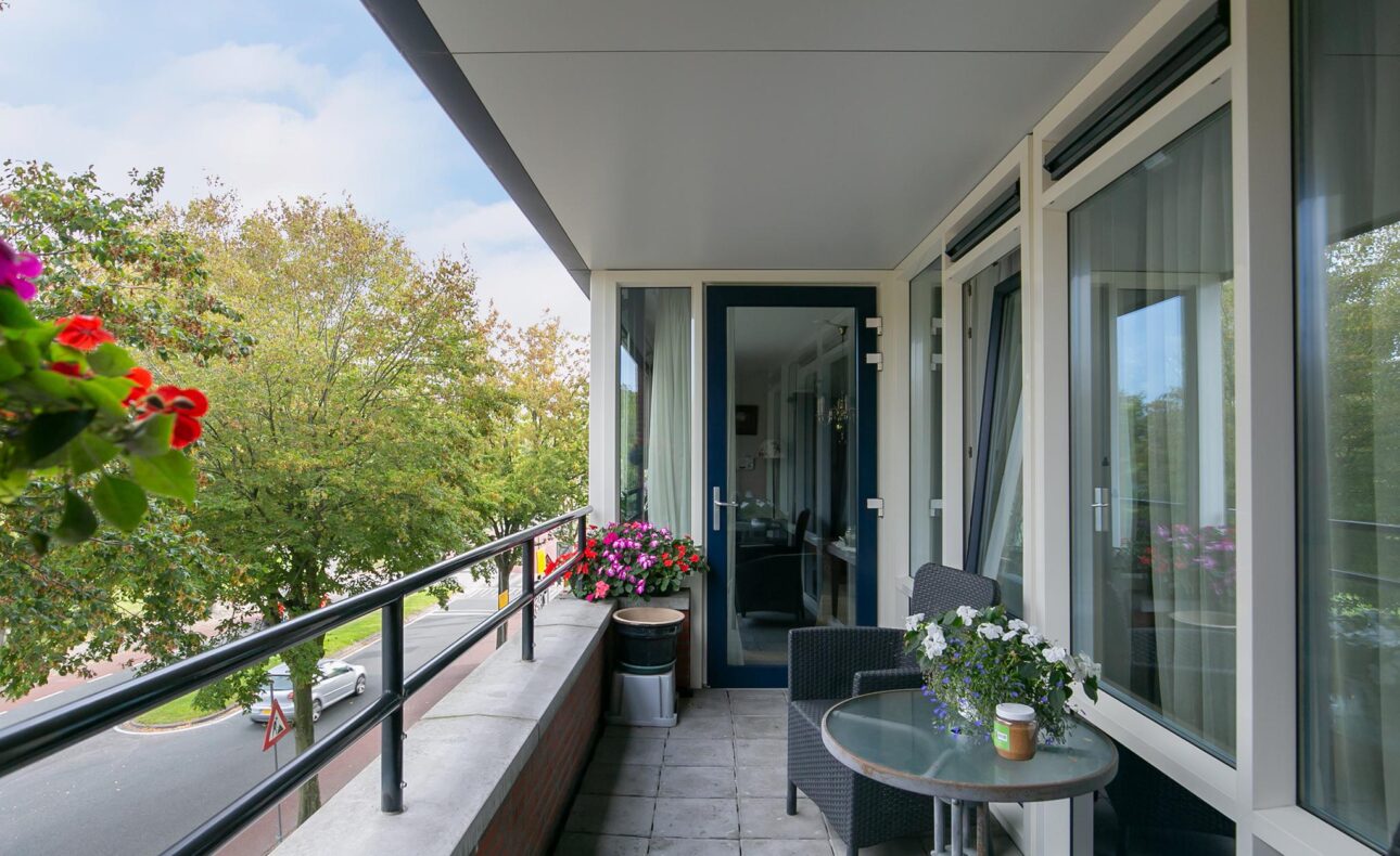 Balkon met een tafel, twee stoelen en bloemen, met uitzicht op een groene straat en bomen onder een bewolkte hemel.