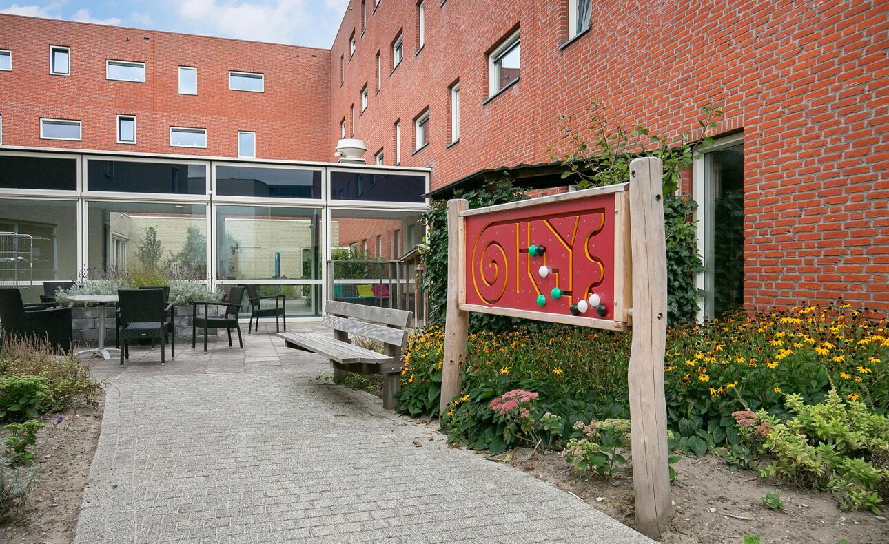 Binnentuin met rode bakstenen muren, een houten bank, kleurrijk bord en bloemen. Grote ramen bieden zicht op het interieur.