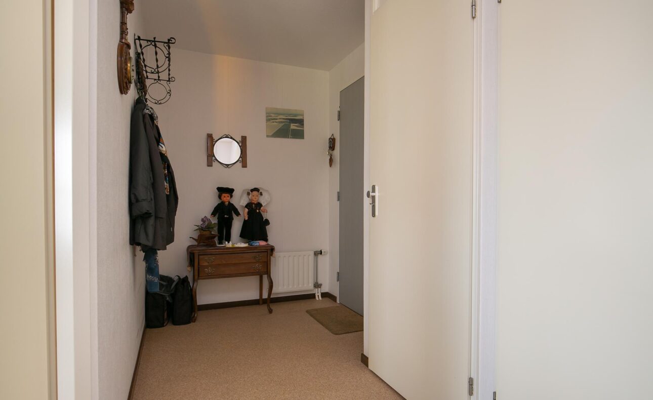 Een smalle hal met een houten tafel, twee poppen, een spiegel aan de muur en jassen aan de kapstok. De vloer is bedekt met tapijt.