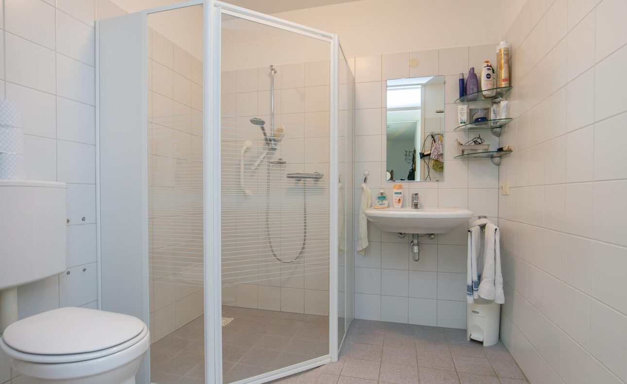 Een moderne badkamer met een glazen douchecabine, wastafel met spiegel, toilet en witte tegels op de muren en vloer.