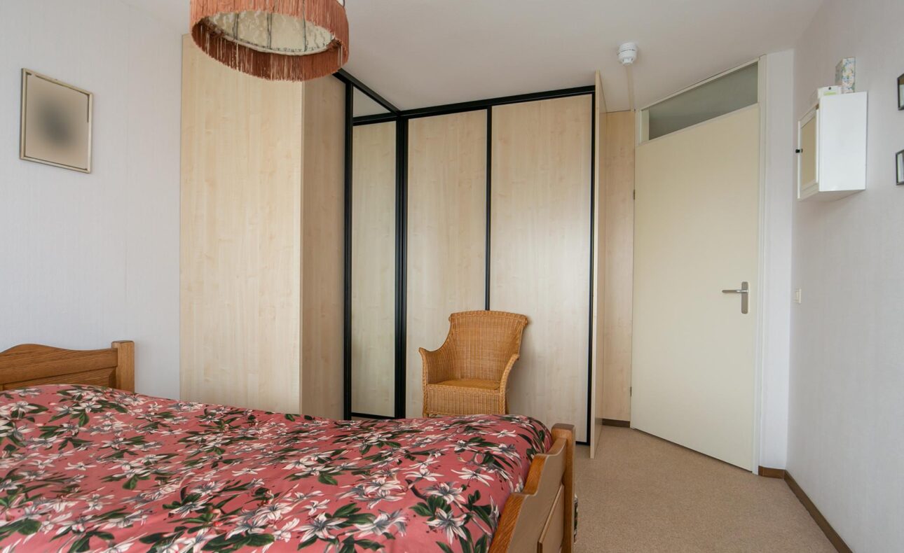 Slaapkamer met een houten bed met bloemenbedekking, een rieten stoel, en een grote kast met spiegeldeuren naast een witte deur.