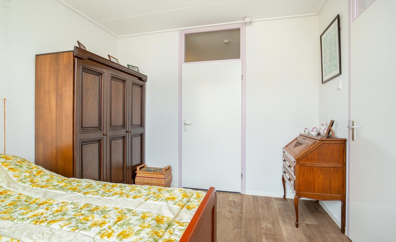 Slaapkamer met houten kast, bureau en bed met geelgroen bloemenpatroon. Witte muren en houten vloer. Rustige, eenvoudige inrichting.