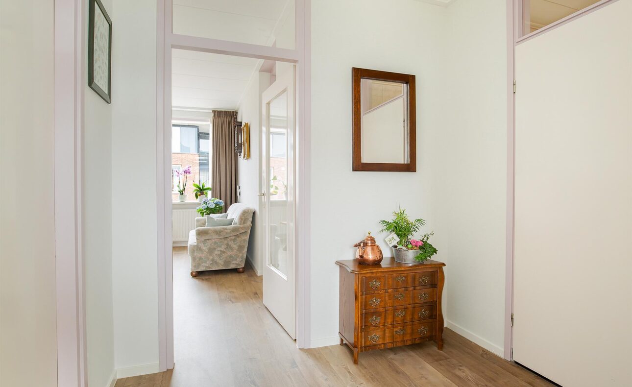 Hal met houten vloer en een antiek kastje met planten. Een spiegel hangt aan de muur. Zicht op een lichte woonkamer.