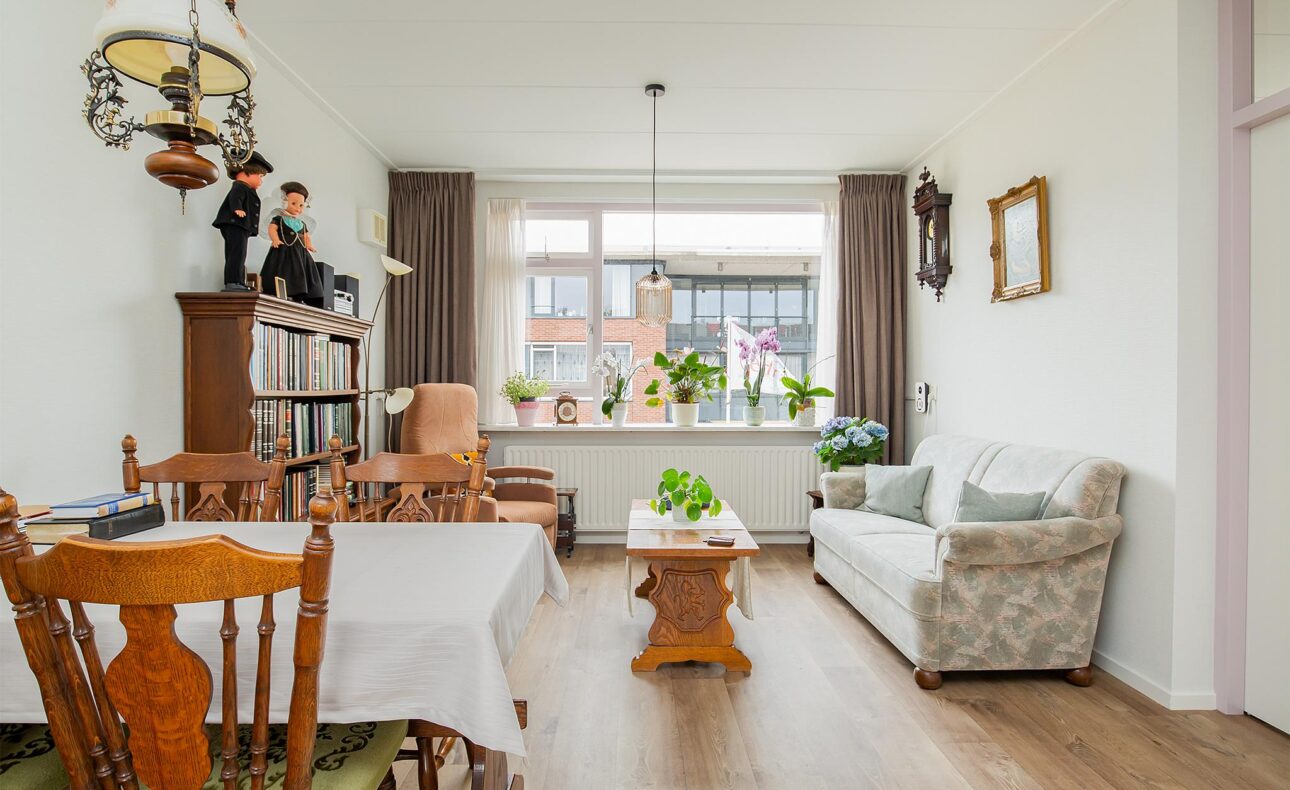 Woonkamer met houten meubels, een bank, planten en een grote raam met uitzicht op een gebouw. Gezellige en lichte sfeer.