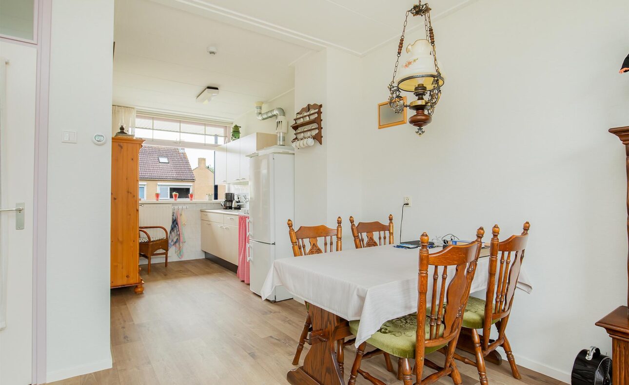 Eetkamer met houten tafel en stoelen, witte muren, en een hanglamp. Achtergrond toont een keuken met raam en kast.