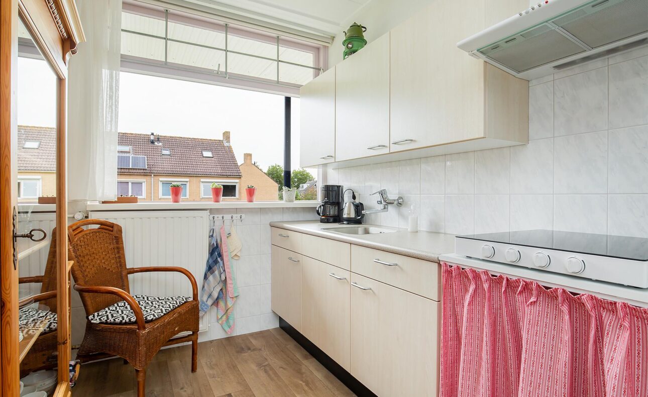 Keuken met witte kasten, een fornuis, en een rieten stoel. Roze gordijn onder het aanrecht en uitzicht op huizen buiten.