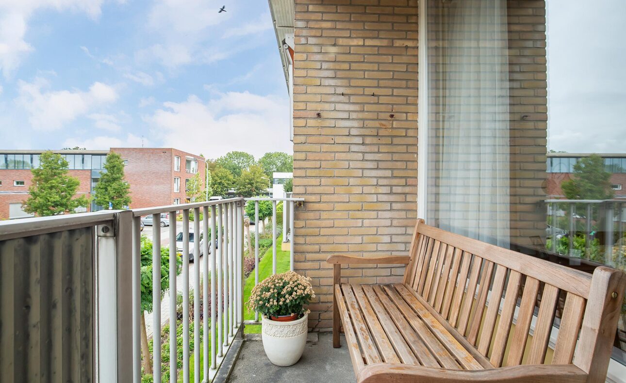 Balkon met houten bank en bloempot met bloemen, uitzicht op een rustige straat met bomen en gebouwen onder een blauwe lucht.