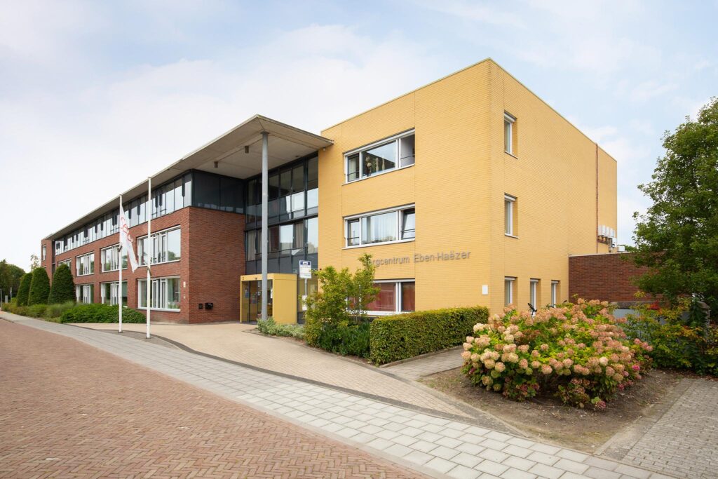Een modern gebouw met gele en rode bakstenen, omgeven door groen en bloemen, onder een heldere lucht. Het heeft meerdere verdiepingen.