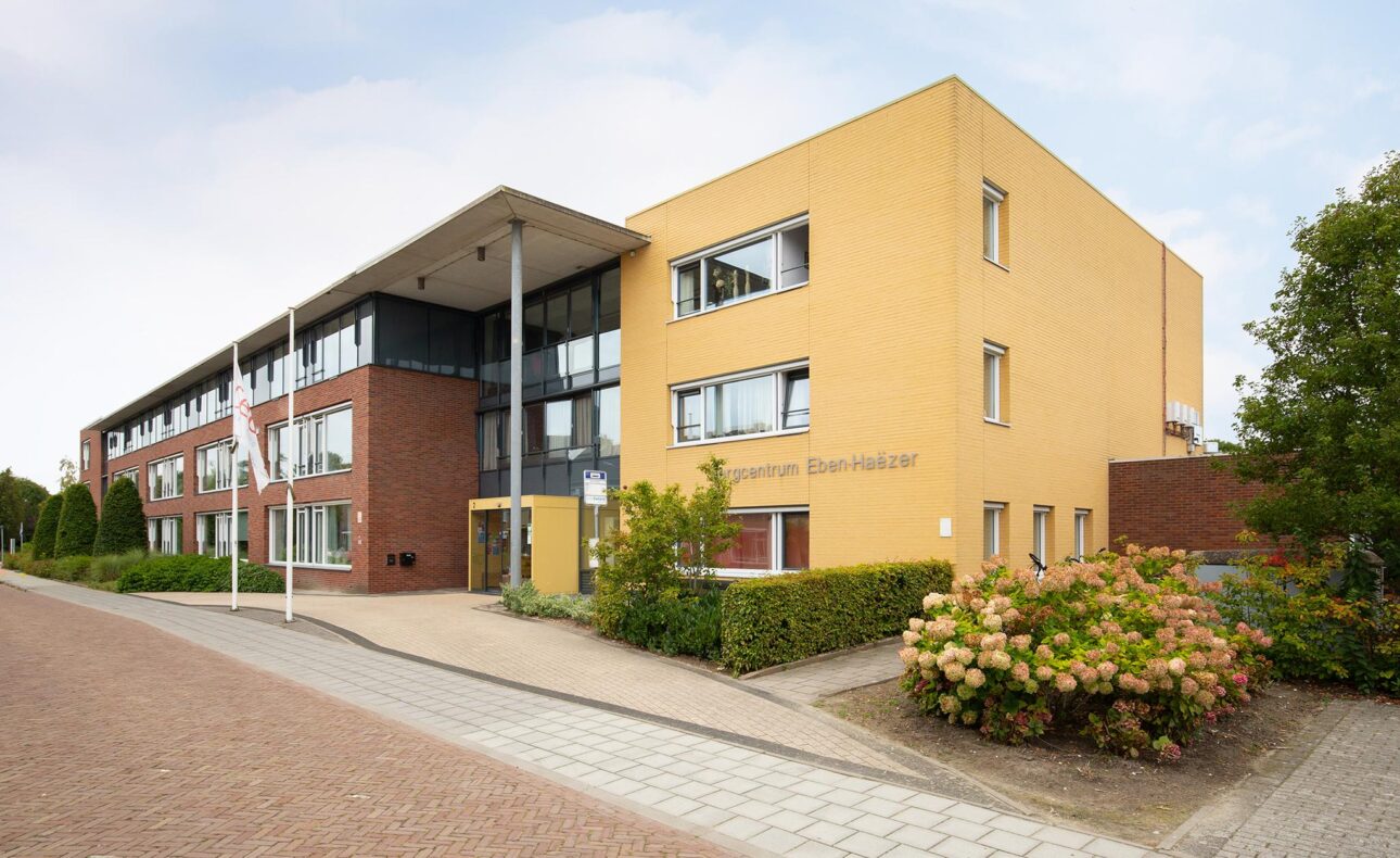 Een modern gebouw met gele en rode bakstenen, omgeven door groen en bloemen, onder een heldere lucht. Het heeft meerdere verdiepingen.