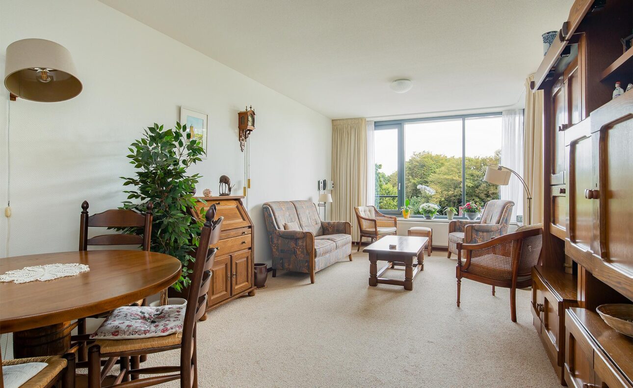 Woonkamer met houten meubels, een ronde eettafel, planten en een groot raam met uitzicht op een groene tuin.