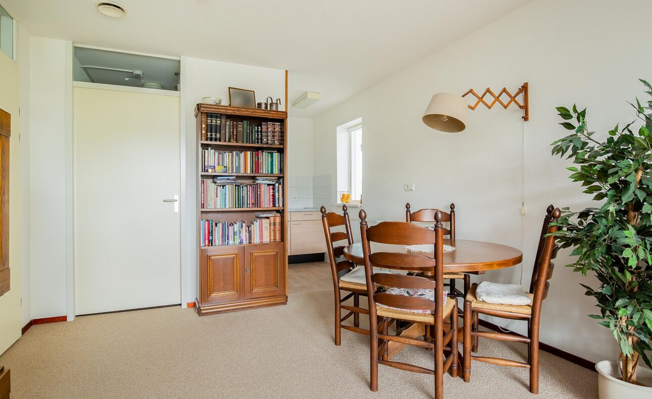 Een lichte kamer met een ronde houten tafel en vier stoelen. Links staat een boekenkast en rechts een grote plant.