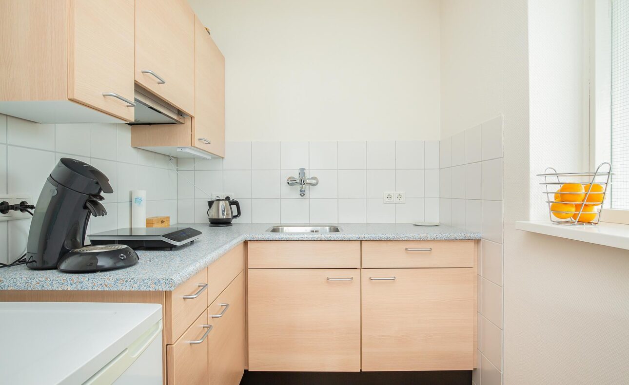 Kleine keuken met houten kasten, een koffiezetapparaat, een waterkoker en een fruitschaal met sinaasappels op de vensterbank.