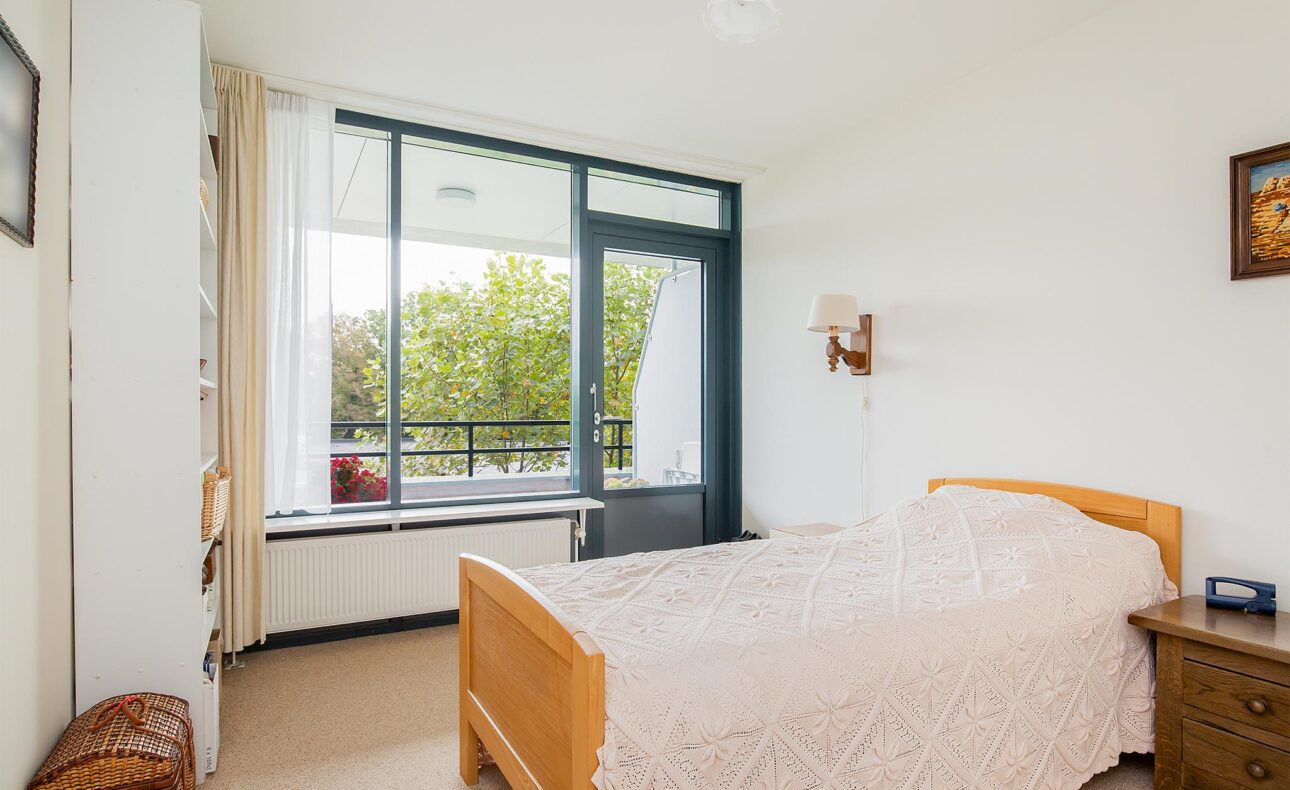 Slaapkamer met een houten bed, wit beddengoed, grote ramen met uitzicht op bomen en een schilderij aan de muur.