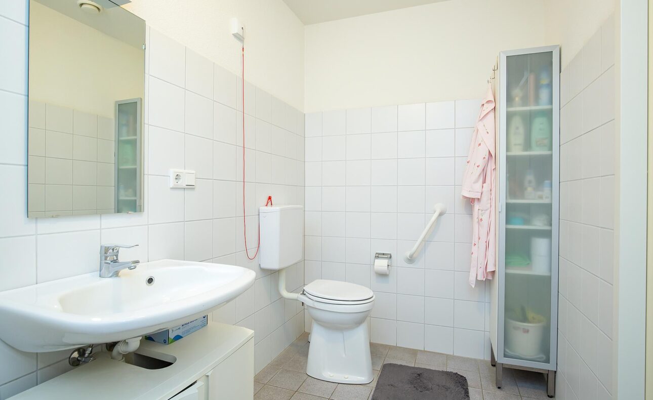 Badkamer met witte tegels, wastafel, spiegel, toilet met steunbeugel, glazen kast en roze badjas aan de muur.