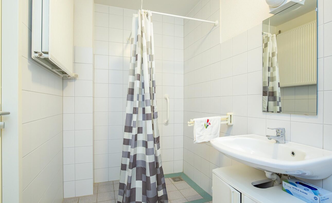 Badkamer met witte tegels, een douchegordijn met grijs-wit patroon, wastafel met spiegel en handdoek aan de muur.