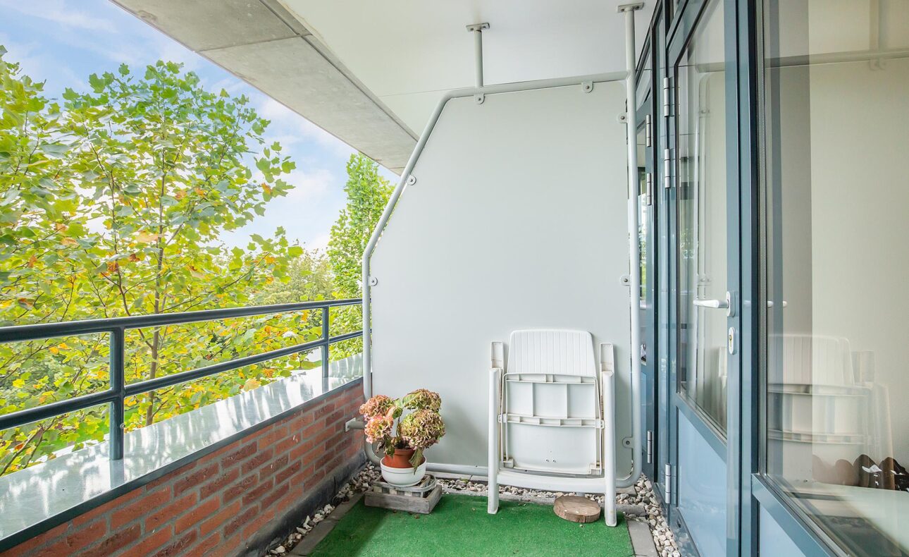 Balkon met groene kunstgrasmat, witte klapstoel en bloempotten. Links een bakstenen muur en uitzicht op groene bomen.