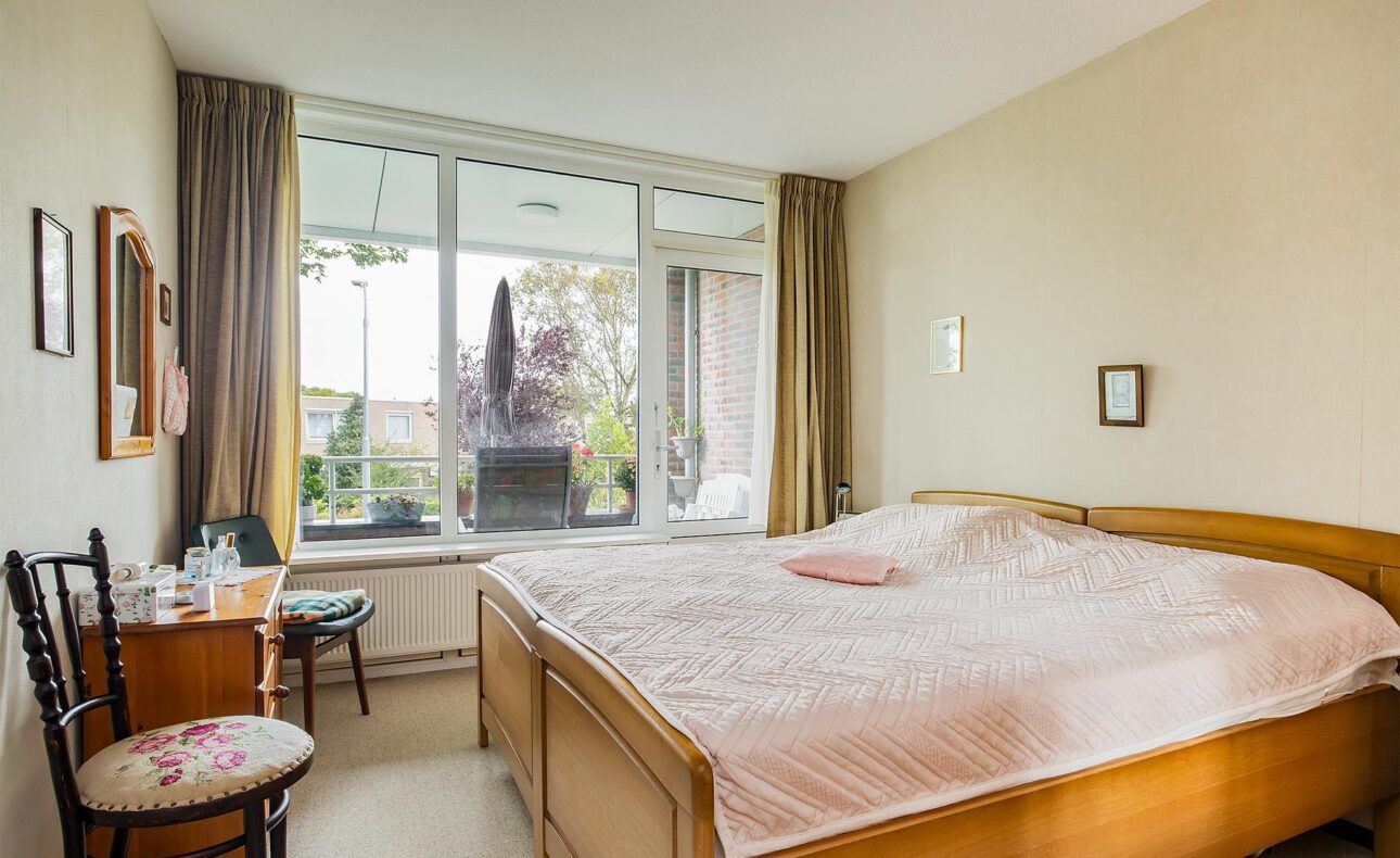 Slaapkamer met een groot bed, houten meubels en een stoel. Er is een raam met uitzicht op een balkon met planten.