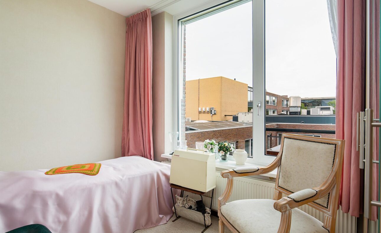 Een lichte kamer met een bed, roze gordijnen, een stoel en een tafel met bloemen, met uitzicht op gebouwen buiten.