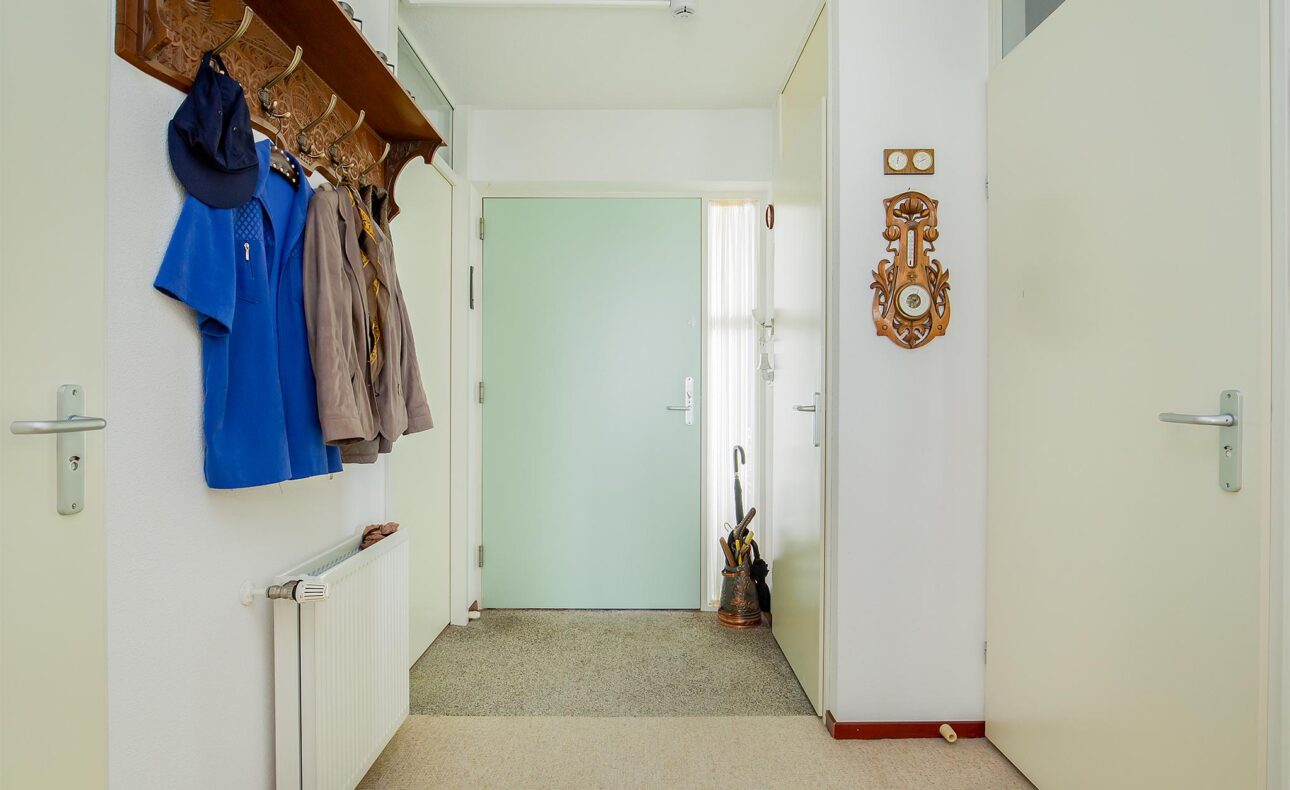 Een gang met een kapstok met jassen en hoeden, een klok aan de muur, en een deur aan het einde naast een paraplu.