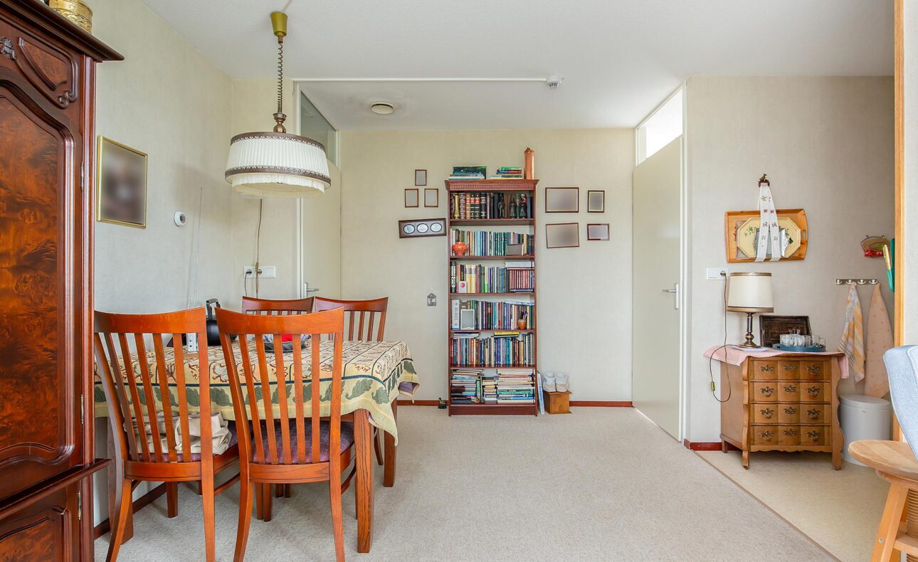 Eetkamer met houten tafel en stoelen, een boekenkast vol boeken, en een vintage lamp. De muren zijn lichtgekleurd.
