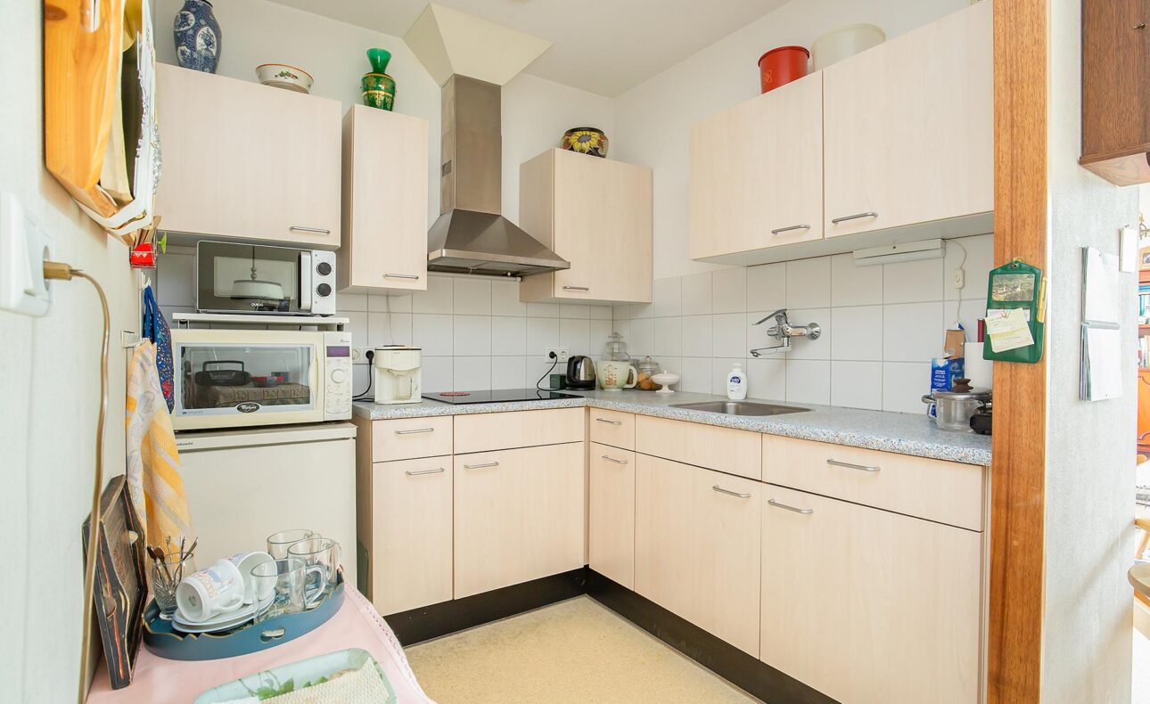 Kleine keuken met lichte houten kasten, een magnetron, fornuis en afzuigkap. Er staan diverse keukenspullen op het aanrecht.