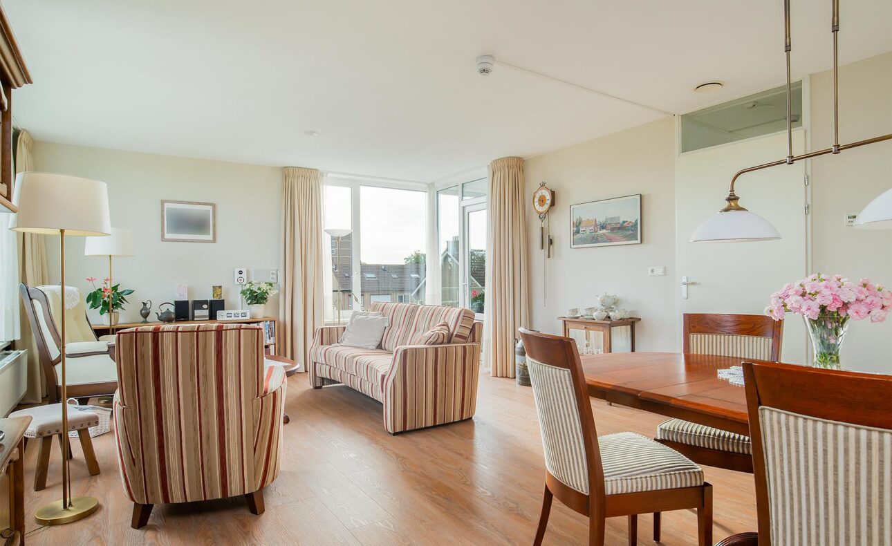 Woonkamer met houten vloer, gestreepte bank en stoelen, eettafel met bloemen, en grote ramen met uitzicht op buiten.