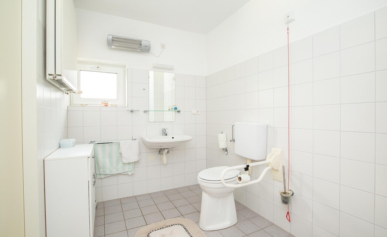 Een lichte, witte badkamer met een toilet, wastafel en spiegel. Er hangt een handdoek en er is een klein raam aanwezig.