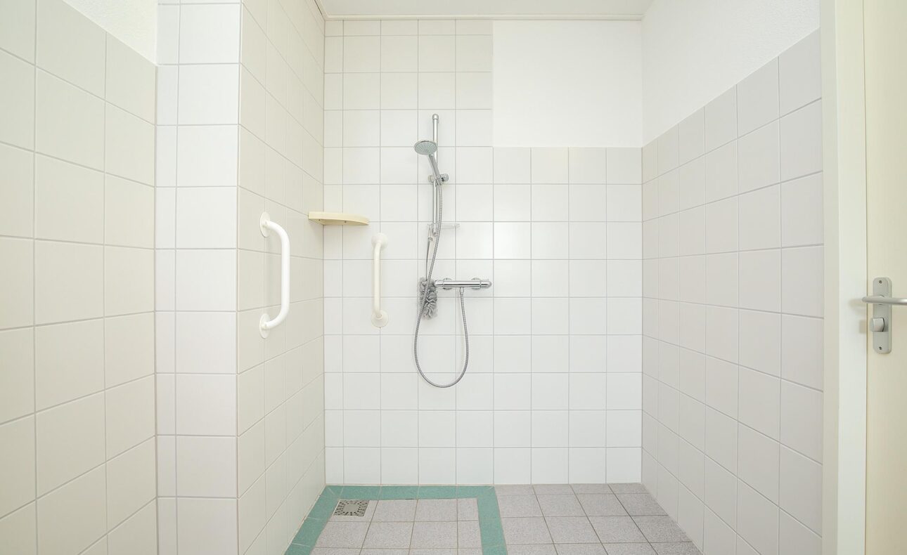 Een ruime, witte badkamer met een inloopdouche, voorzien van een handgreep en een douchekop aan de muur.