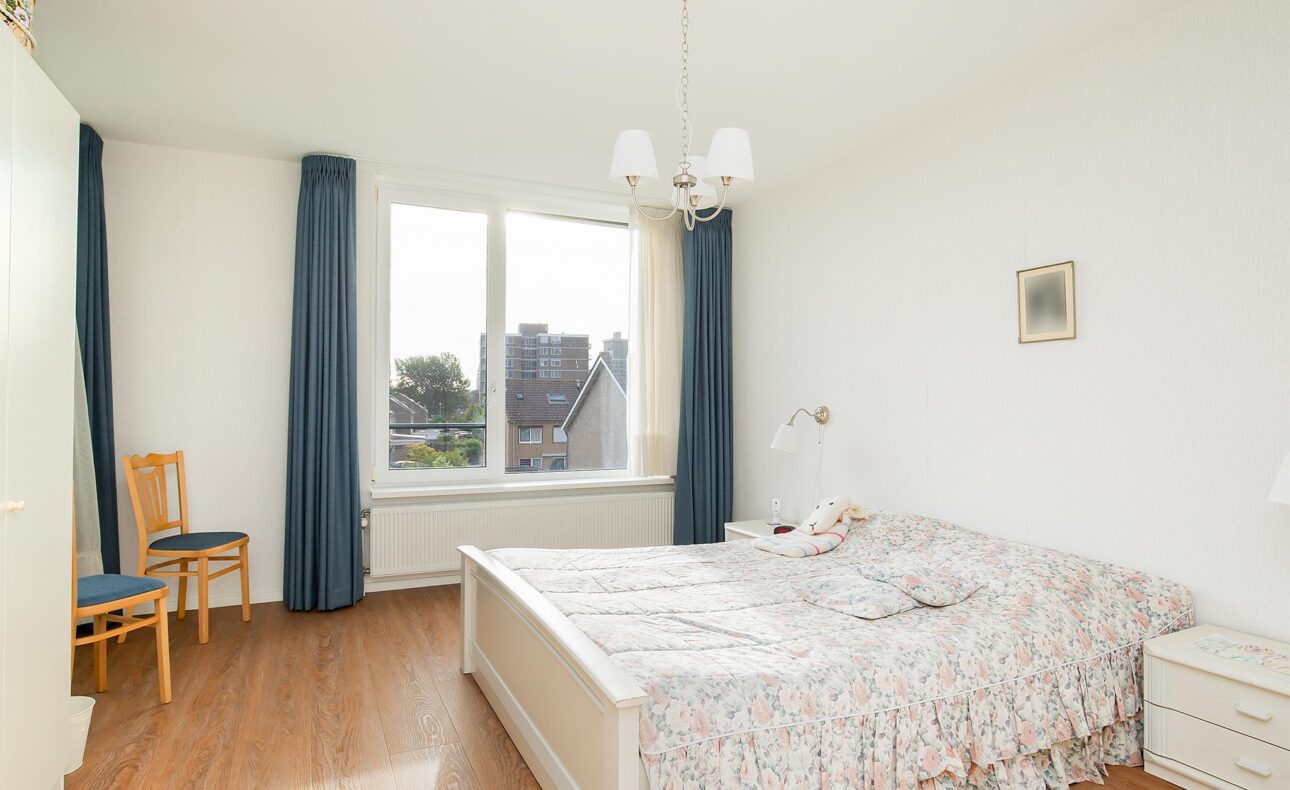 Slaapkamer met een tweepersoonsbed, houten vloer, blauwe gordijnen en een raam met uitzicht op gebouwen en bomen.