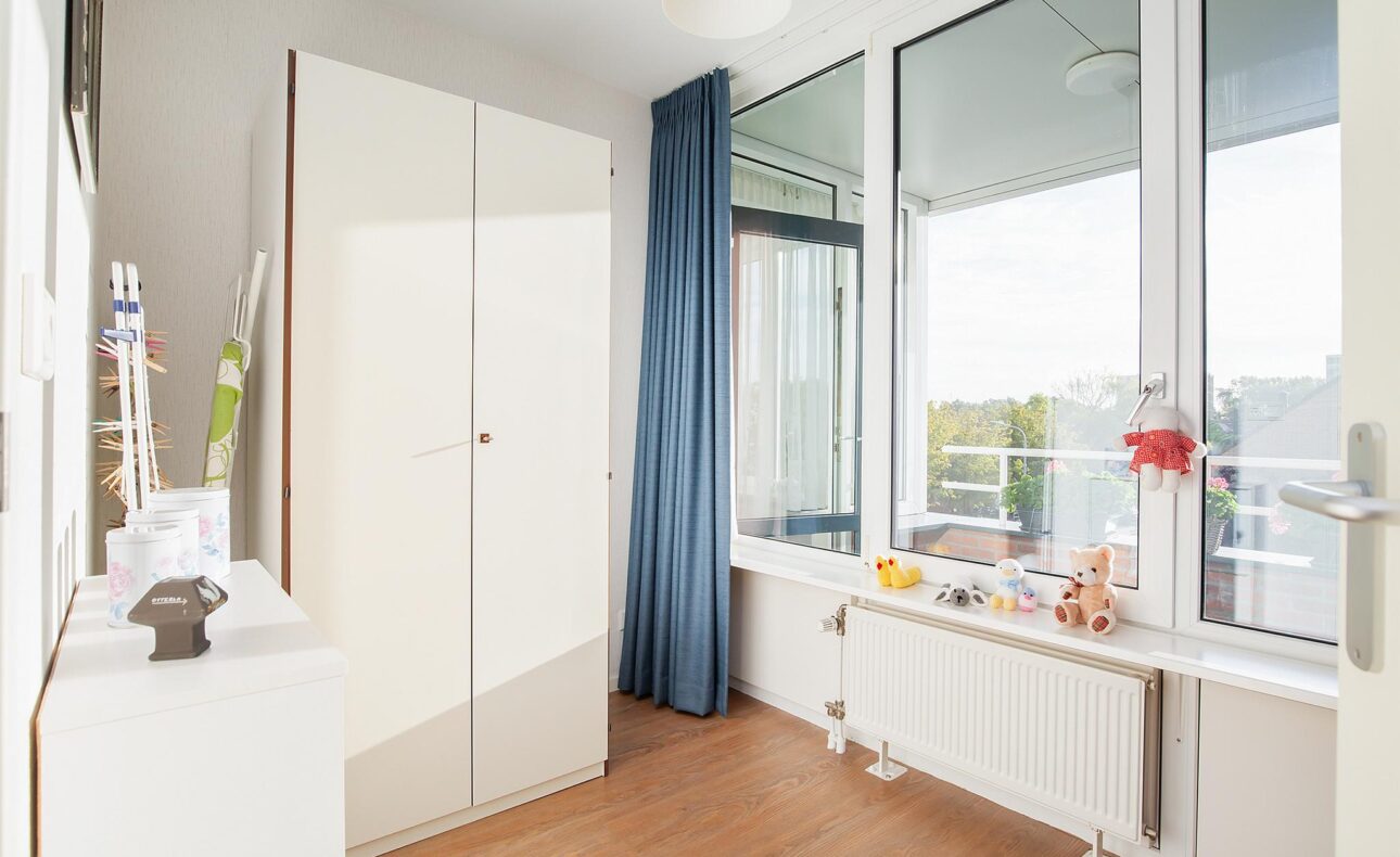 Een lichte kamer met een witte kast, blauwe gordijnen en een groot raam. Er staan decoraties op de vensterbank.
