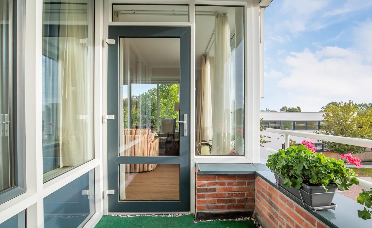 Balkon met groene vloerbedekking, bloembakken met bloemen en een glazen deur naar een lichte kamer met gordijnen.