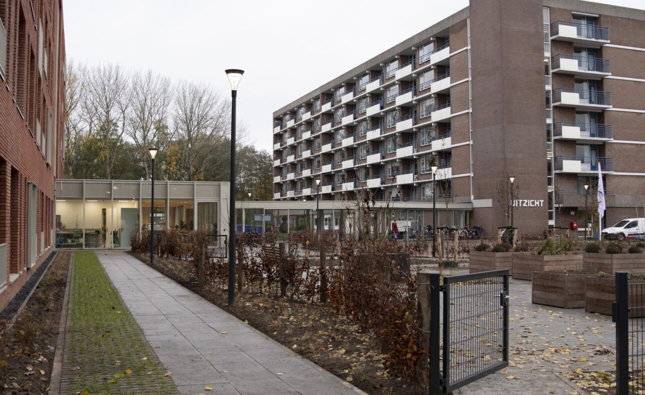 Afbeelding van een modern appartementencomplex met meerdere verdiepingen, omgeven door een tuin en een betegeld pad op een bewolkte dag.