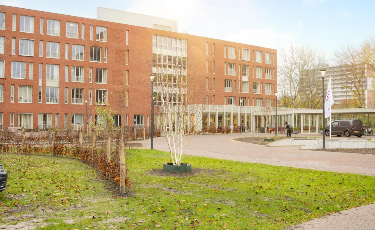 Buitenaanzicht van een modern, rood bakstenen gebouw met meerdere verdiepingen, omgeven door een groen gazon en bomen.