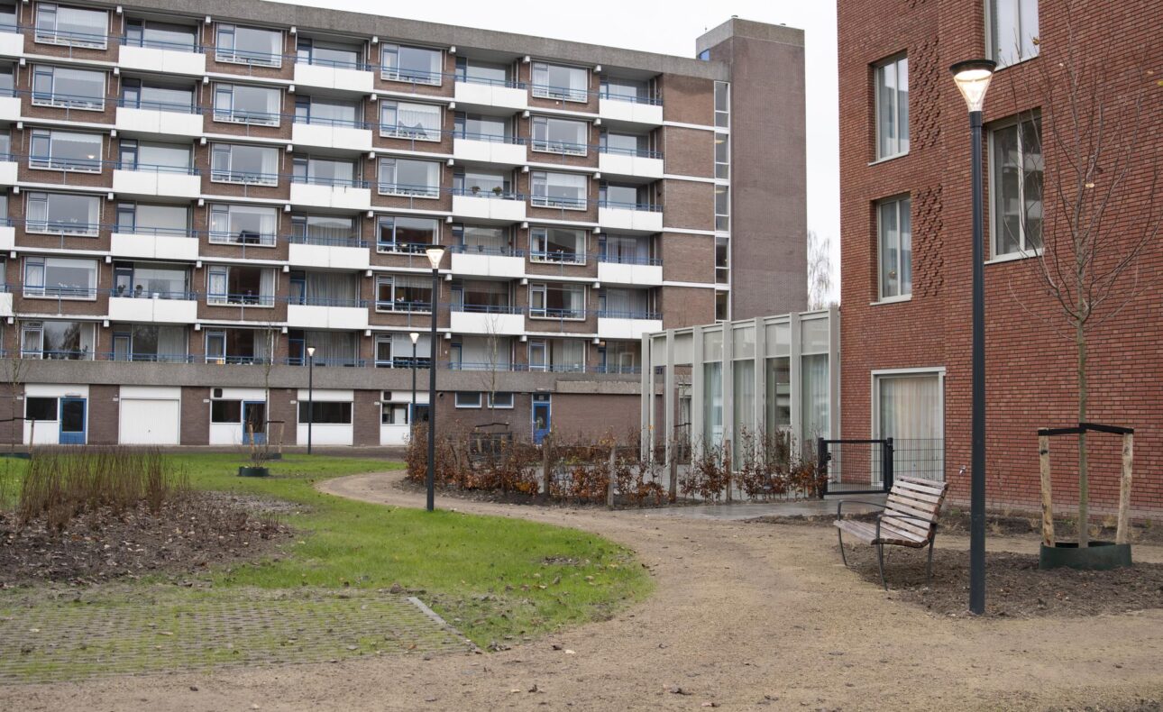 Appartementencomplex met meerdere verdiepingen en balkons, omgeven door een tuinpad en grasveld, naast een bakstenen gebouw.