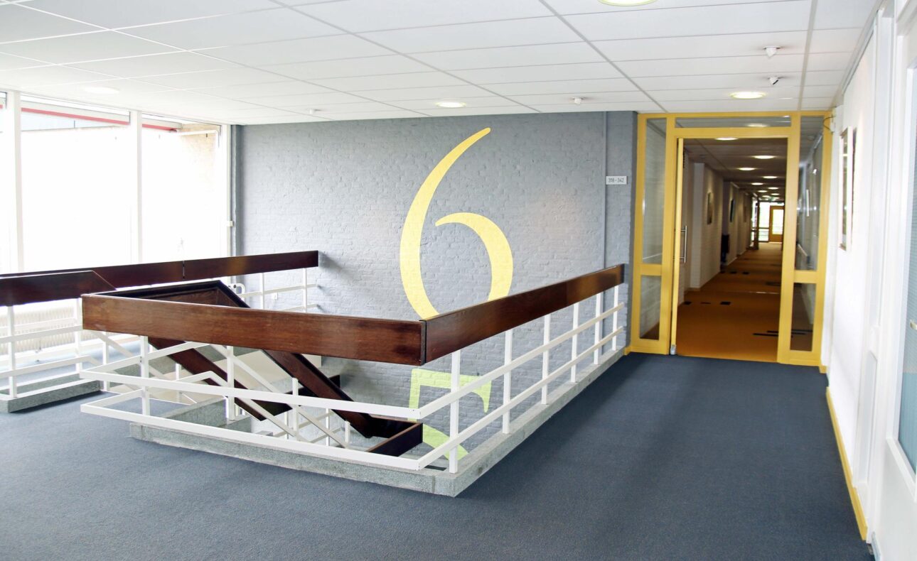 Trap en gang in een gebouw met een groot geel cijfer zes op de muur. De gang heeft een blauwe vloerbedekking en witte muren.