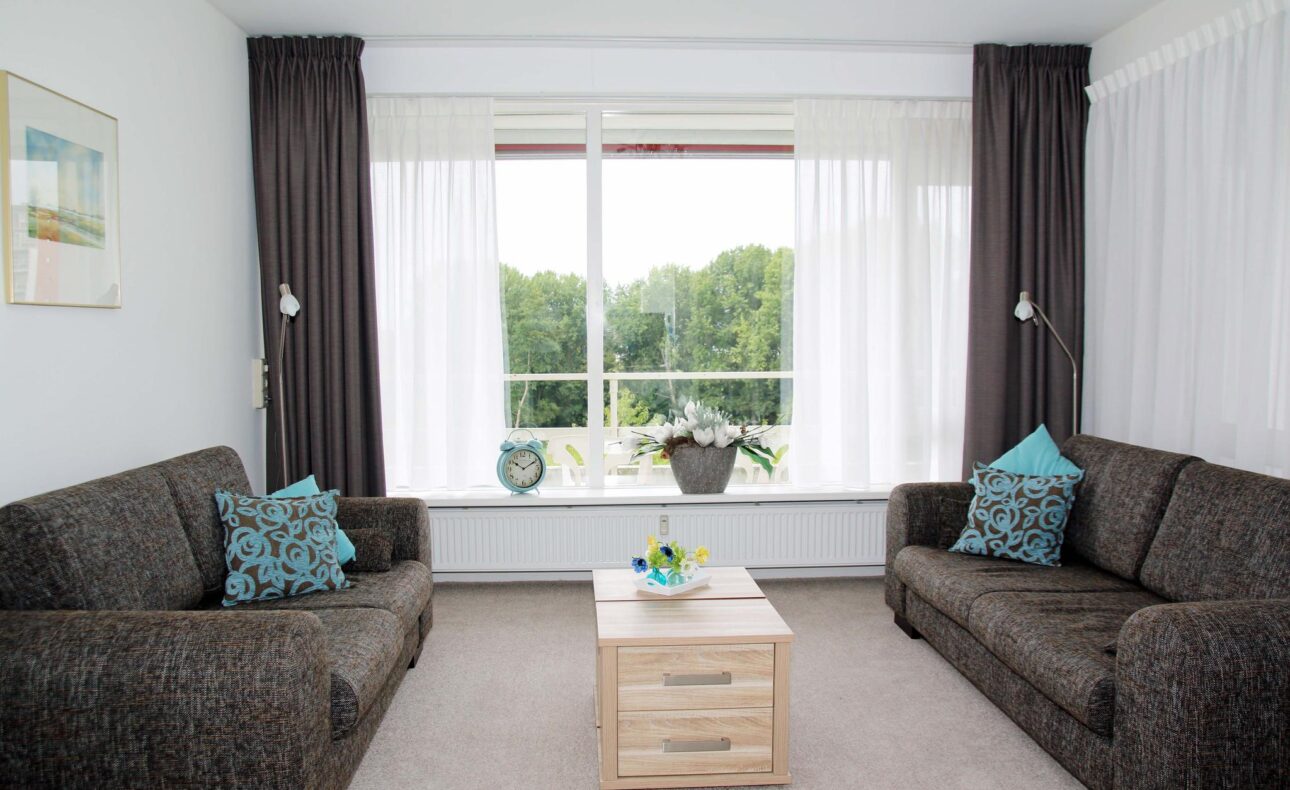 Woonkamer met twee bruine banken, een houten salontafel en een groot raam met uitzicht op groene bomen en witte gordijnen.