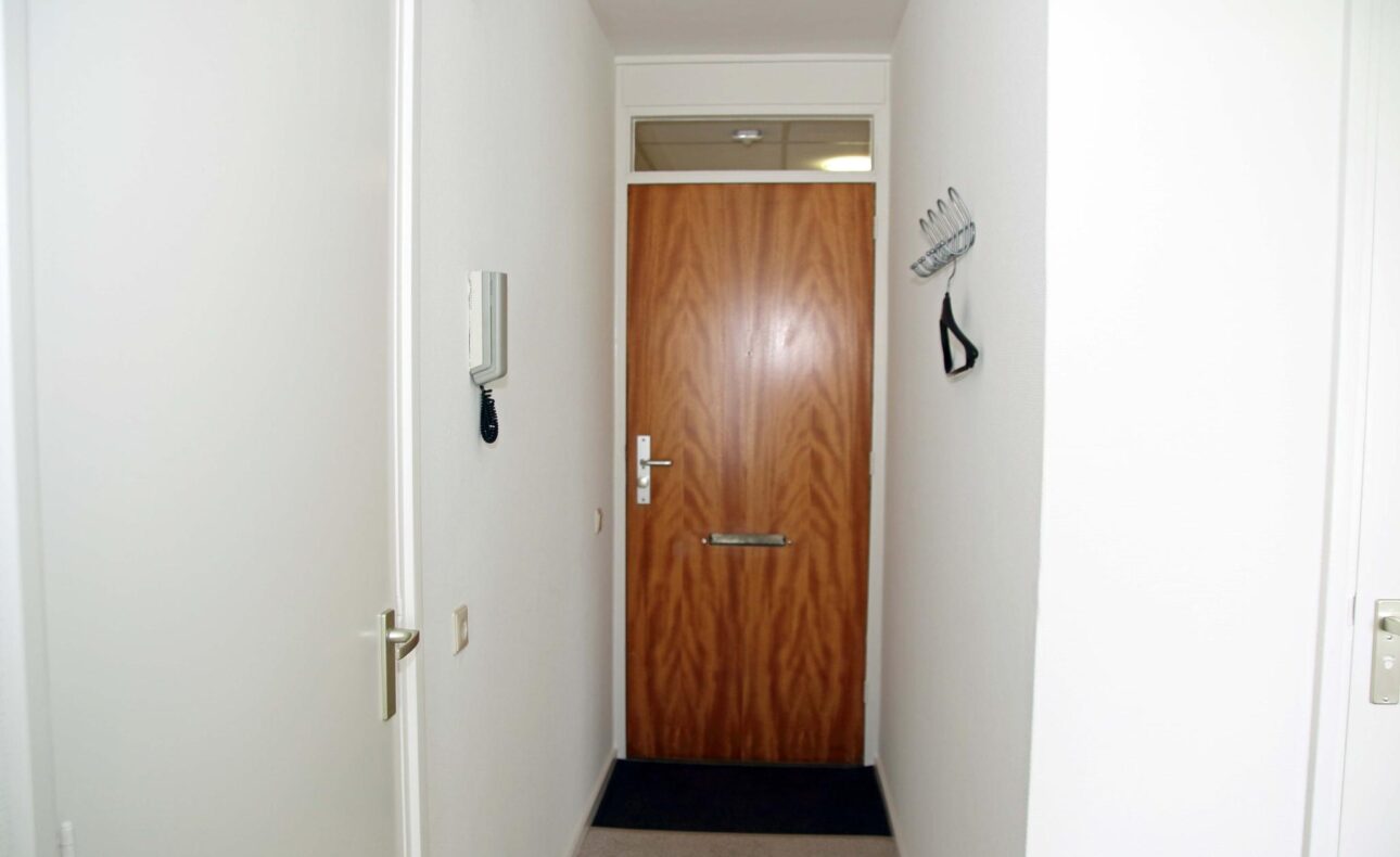 Afbeelding van een smalle gang met witte muren en een houten deur aan het einde. Er hangt een intercom aan de linkermuur.