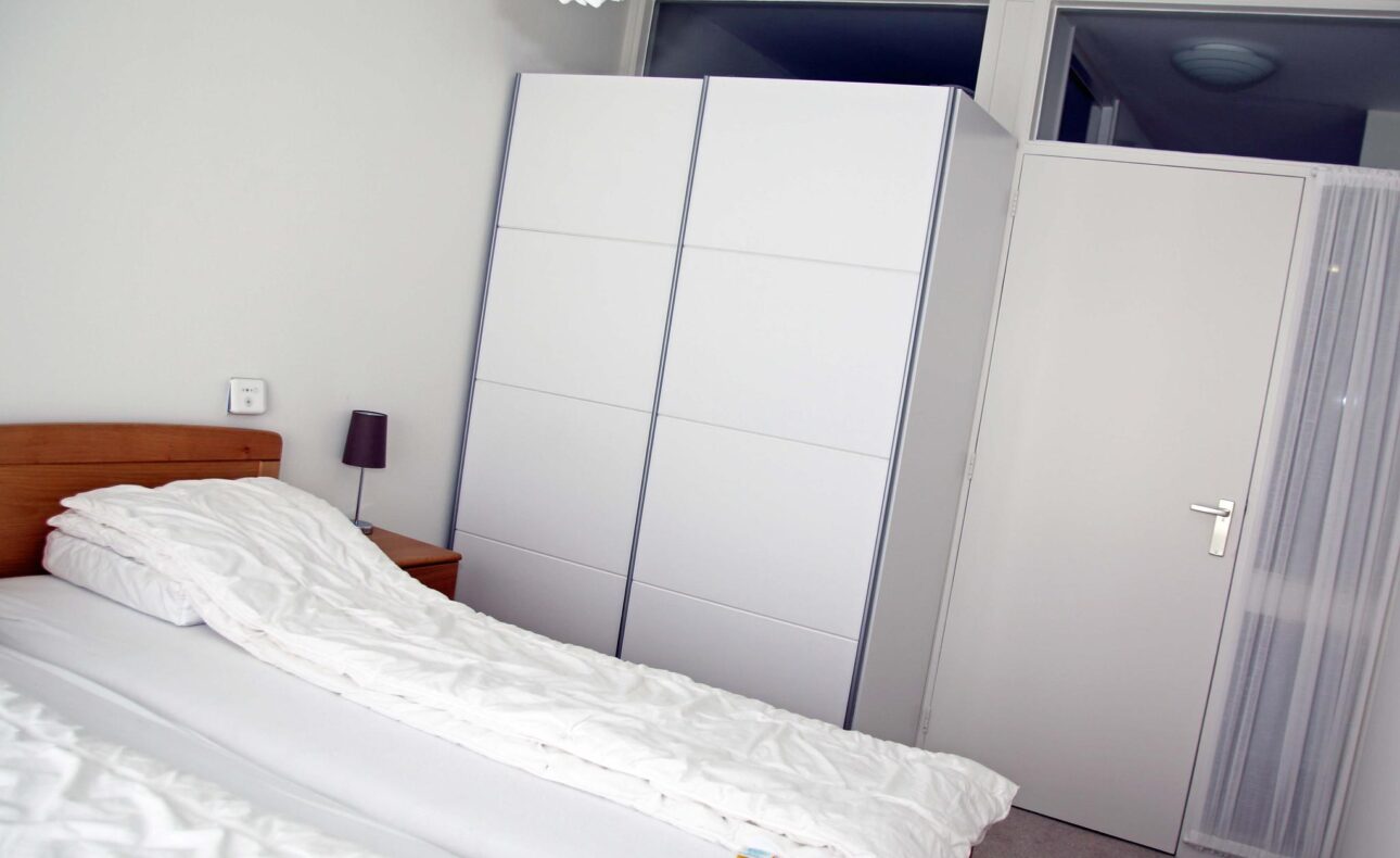 Slaapkamer met een opgemaakt tweepersoonsbed, een witte kledingkast en een deur. Er hangt een lamp aan het plafond.