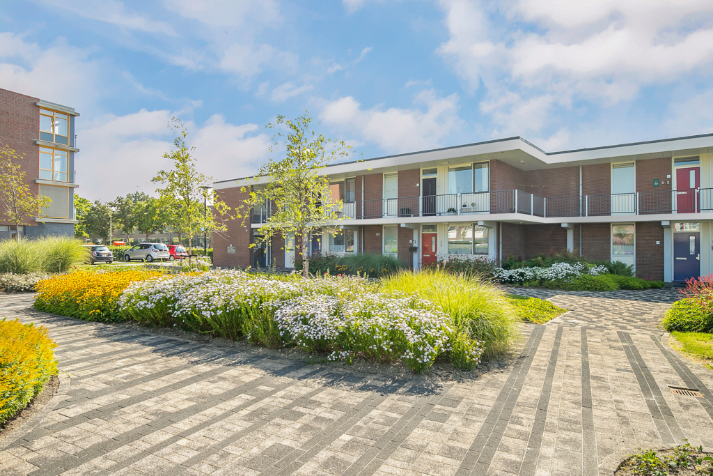 Moderne appartementencomplexen met bakstenen gevels, omgeven door kleurrijke bloemen en groen, onder een blauwe lucht met wolken.
