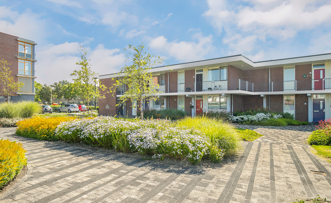 Moderne appartementencomplexen met bakstenen gevels, omgeven door kleurrijke bloemen en groen, onder een blauwe lucht met wolken.