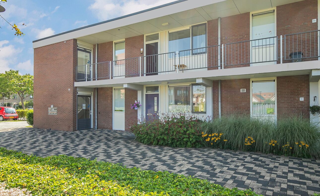 Twee verdiepingen tellend bakstenen gebouw met balkons, omgeven door groen en bloemen, onder een blauwe lucht met wolken.