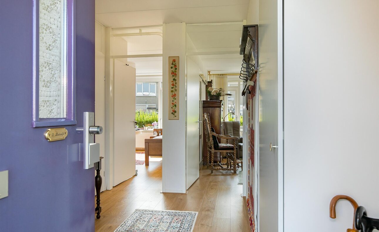 Een open deur geeft zicht op een lichte woonkamer met houten vloer, een tapijt en meubels. Planten staan bij het raam.