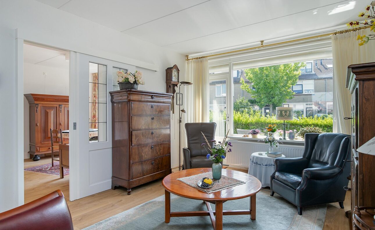 Woonkamer met houten meubels, een ronde tafel met bloemenvaas, leren stoelen en een groot raam met uitzicht op de tuin.