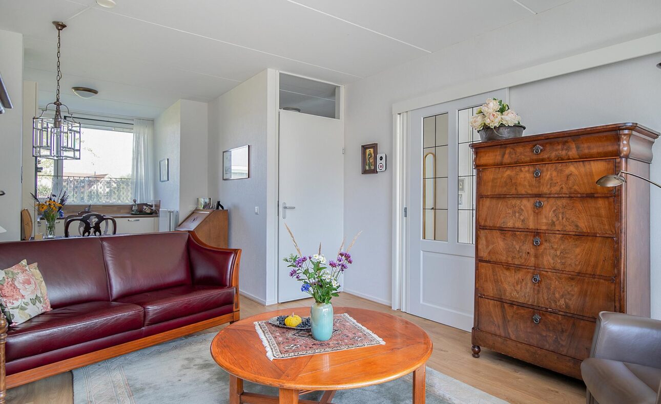 Woonkamer met een bruine leren bank, houten salontafel met bloemenvaas, antieke kast en zicht op de eetkamer en keuken.
