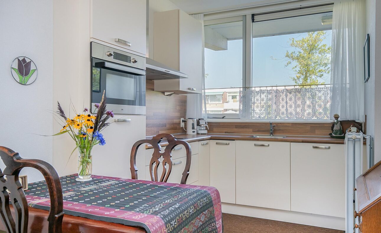 Keuken met witte kasten, een oven, en een tafel met bloemen in een vaas. Groot raam met uitzicht op een boom en gebouwen.